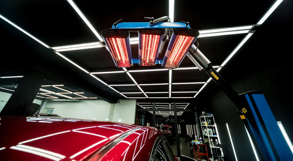 Estúdio de estética automotiva com teto de barras de luz e lâmpadas de secagem sobre um carro vermelho.