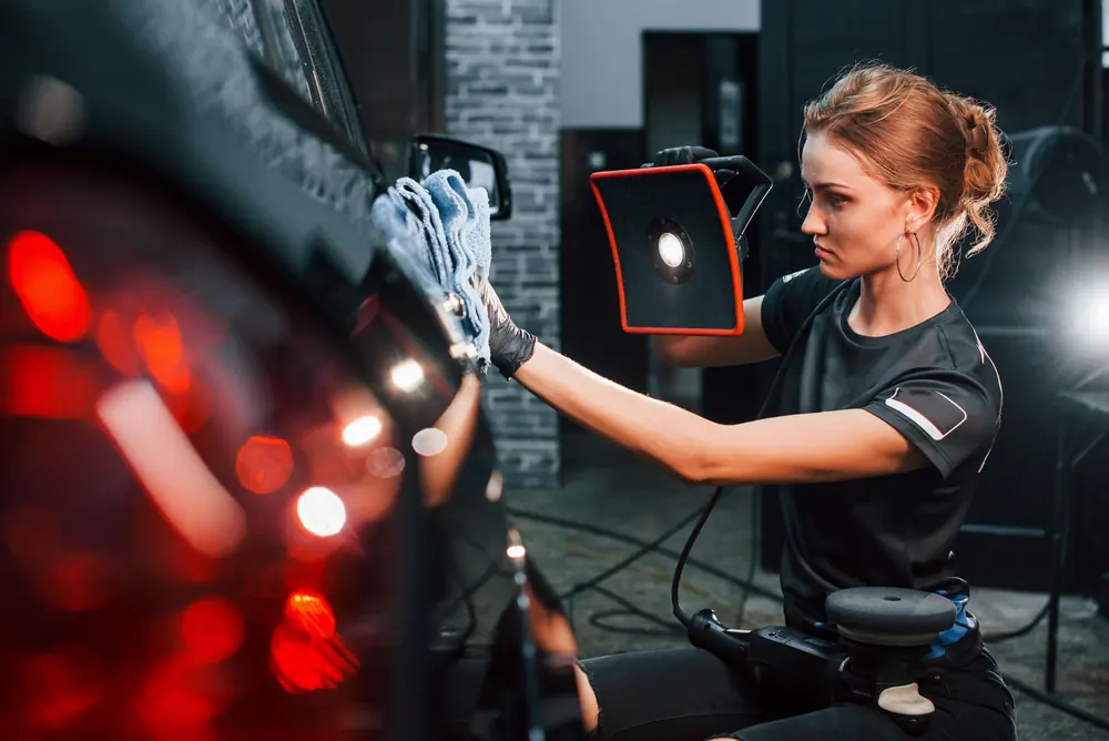 Profissional examinando a lateral de um carro escuro com uma luz de inspeção na mão.