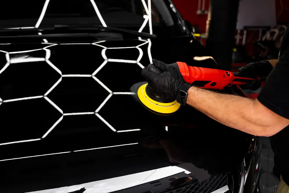Mão de luva preta polindo o capô de um carro preto com uma politriz; o teto de luzes hexagonais aparece refletido na pintura.